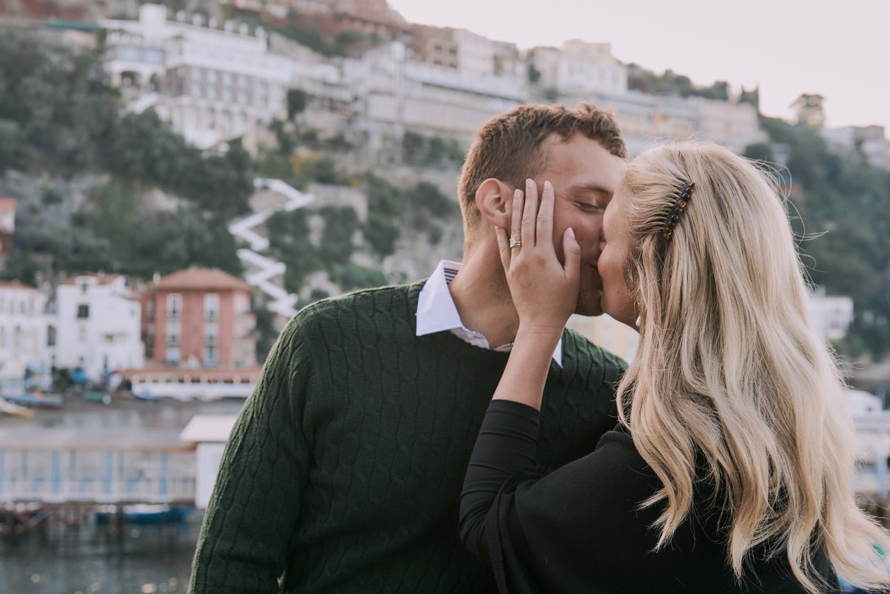 Couple kissing at the Sorrento Marina with colorful waterfront buildings and hillside architecture behind them