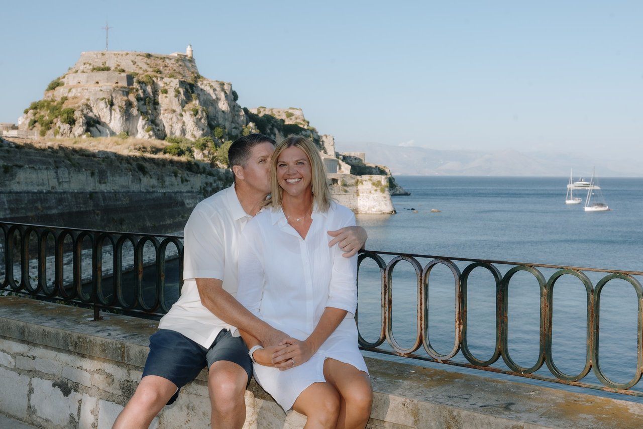 Couple posing by seaside railing with historic clifftop fortress and sailboat in background during daytime