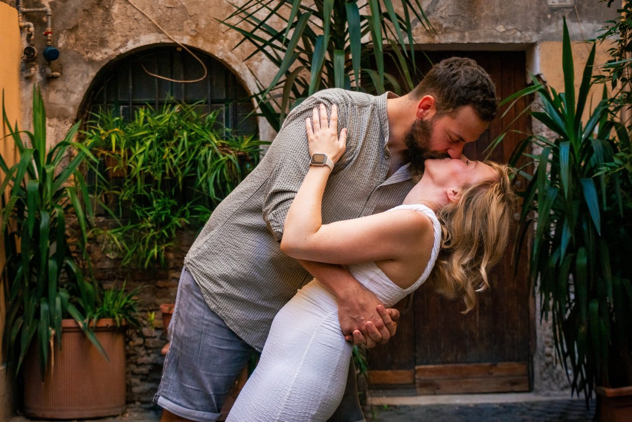 Couple kissing in a lush courtyard surrounded by potted green plants and rustic terracotta walls in Rome