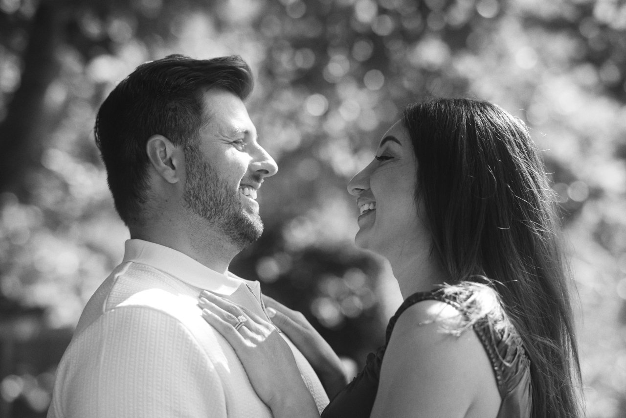 Couple smiling at each other in dappled sunlight beneath trees in black and white