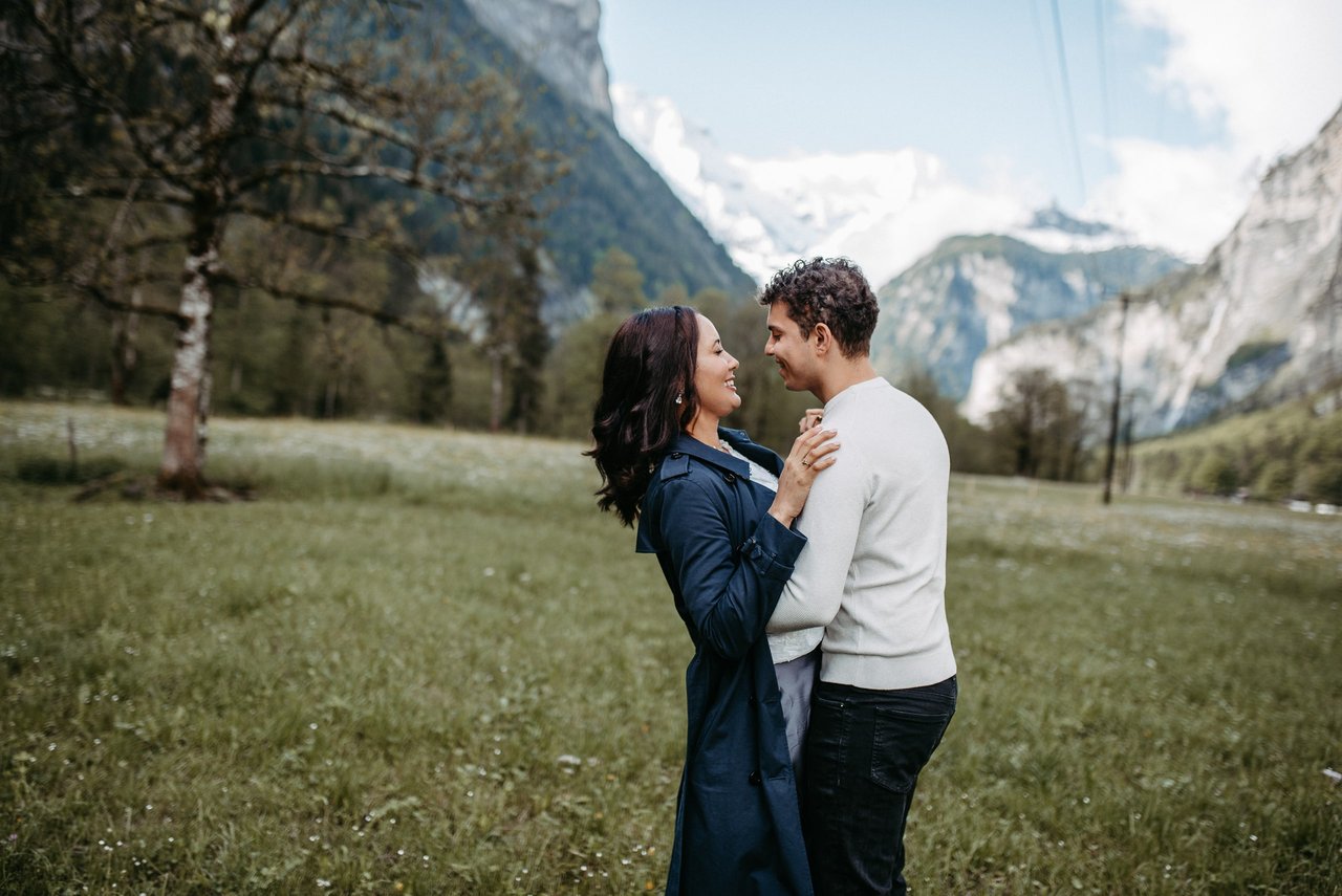 Couple embracing in alpine meadow with snow-capped mountains and valley in background during daytime