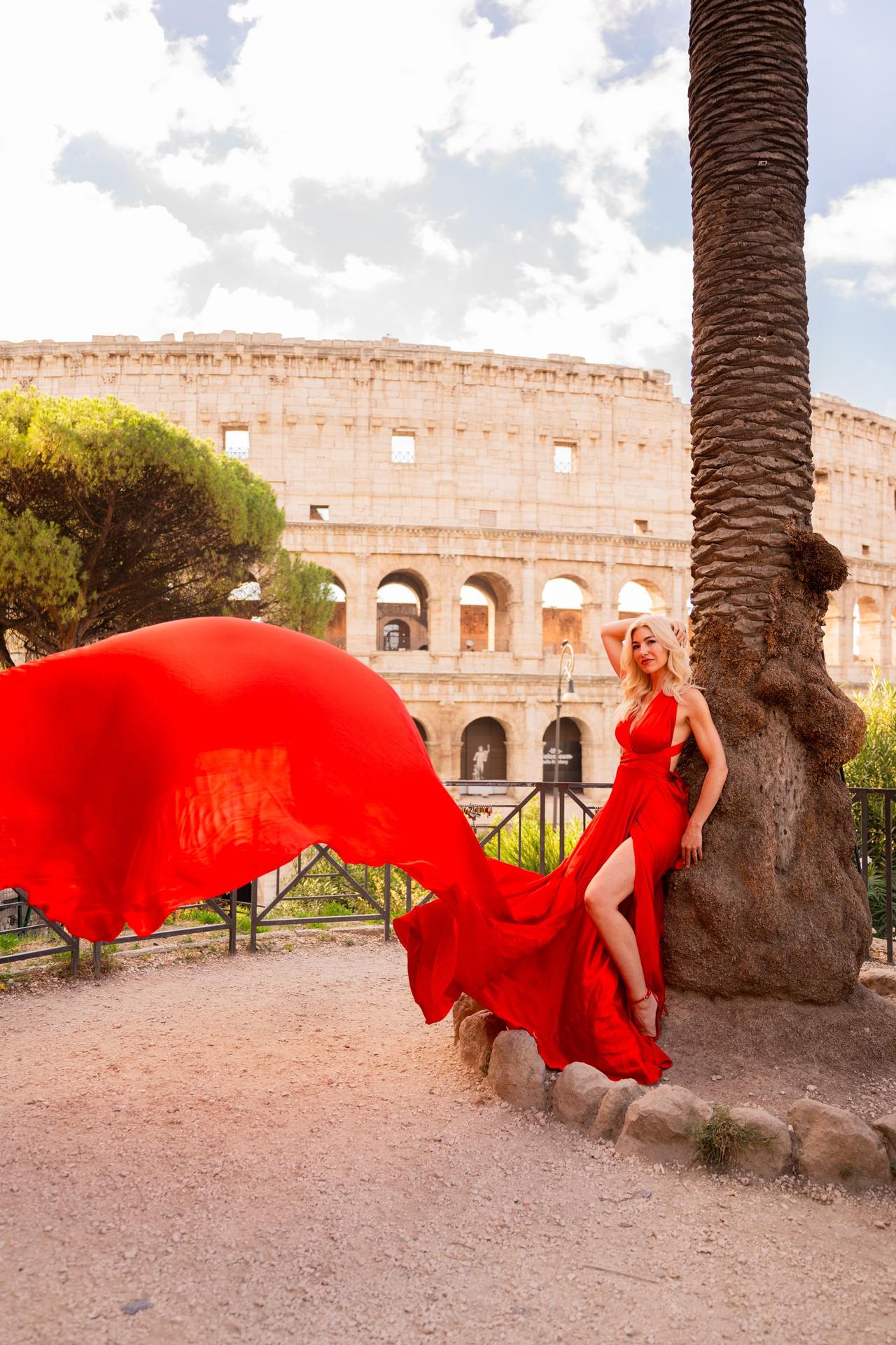 Blonde woman in a billowing red gown leaning against a palm beside the Colosseum in Rome
