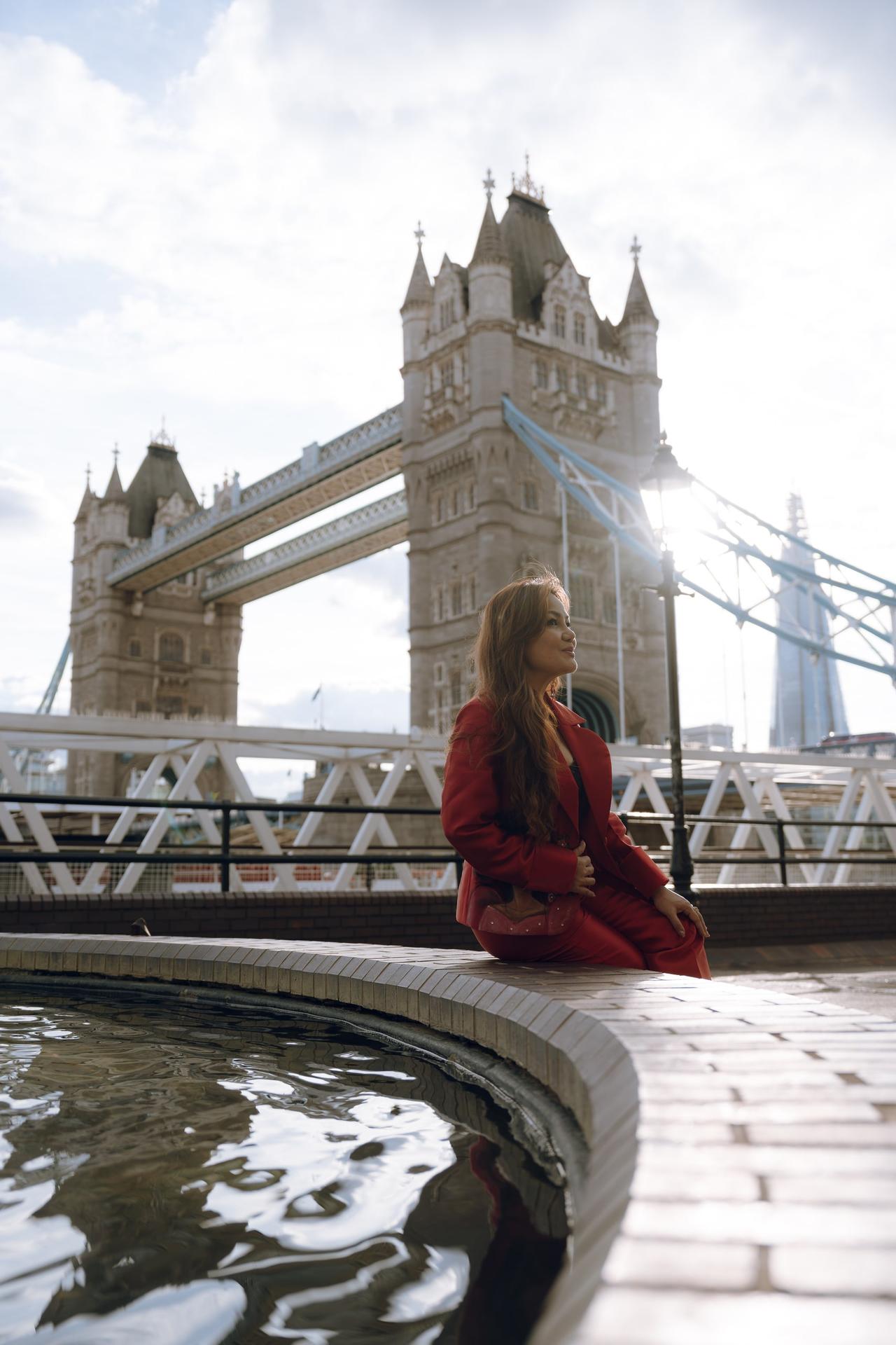 Woman in red coat posing by the Thames with Tower Bridge behind her on a cloudy day