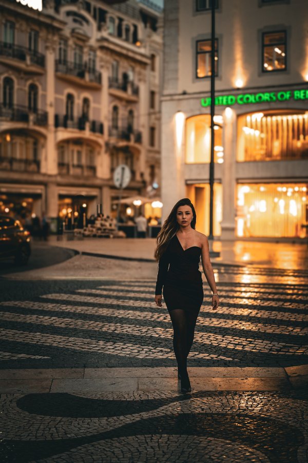 Woman in black dress walking through illuminated Cascais old town street at evening with historic architecture and storefronts