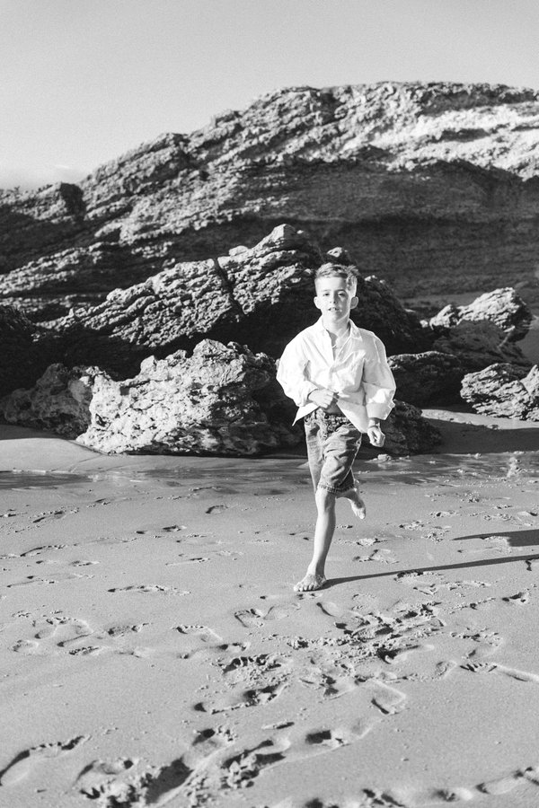 Young boy running barefoot on sandy beach with dramatic rocky cliffs and sea stacks in background, black and white portrait