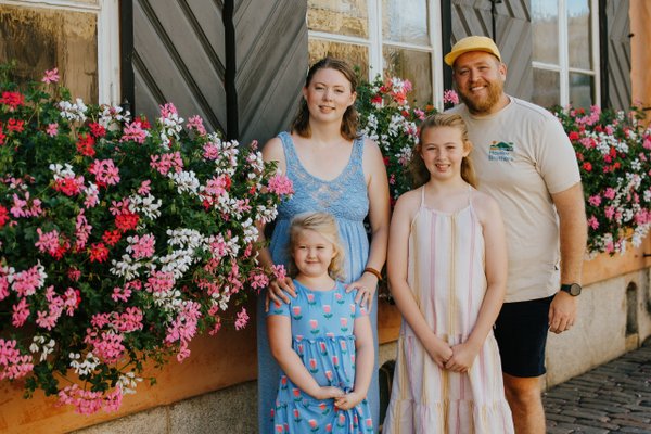 Family of four smiling beside vibrant pink and red flowers against a rustic building in Helsinki city center