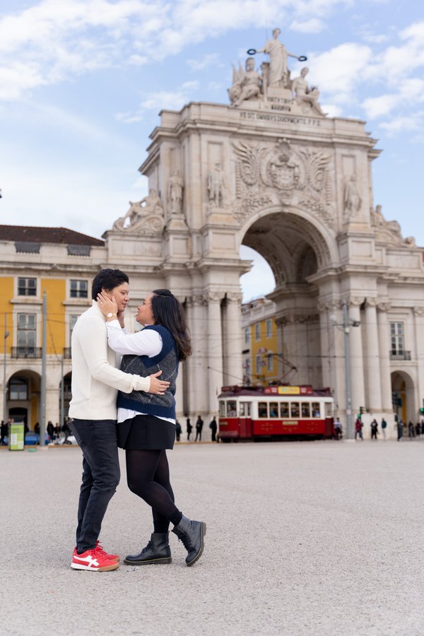 Couple posing together at Praça do Comércio with the Rua Augusta Arch and red tram behind them on a sunny day