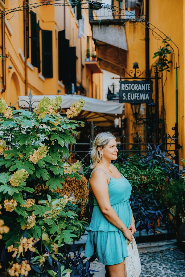 Solo traveler in blue dress among flowering vines on charming yellow-walled Italian street at golden hour