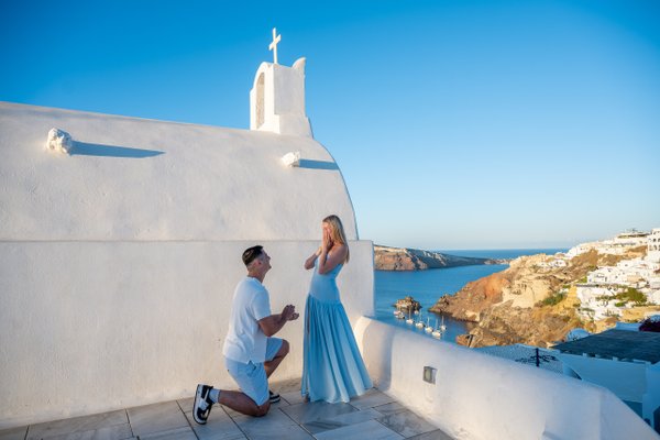 Man kneeling to propose to woman in blue dress at white church in Santorini, overlooking caldera at sunset