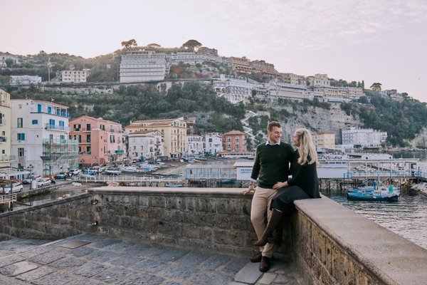 Couple sitting on waterfront wall overlooking colorful buildings and boats in Sorrento Marina during golden hour