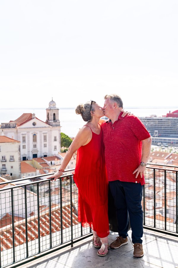 Couple kissing on a sunny rooftop terrace overlooking Alfama's white church and red-tiled roofs in Lisbon