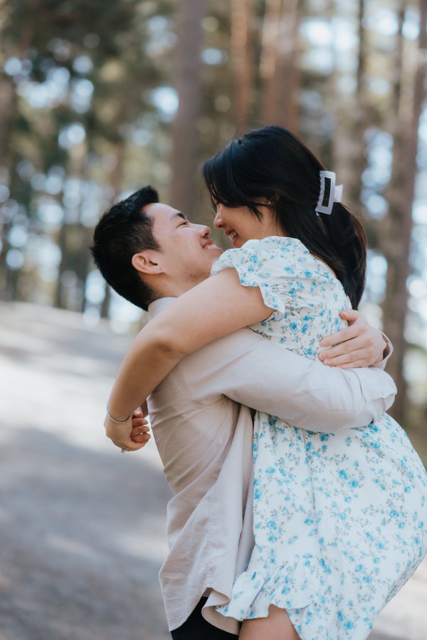 Couple embracing and laughing in a sunlit forest with soft bokeh and tall trees in background