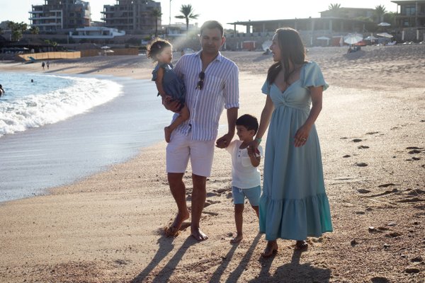 Family of four standing on sandy beach at golden hour with coastal buildings and palm trees in background