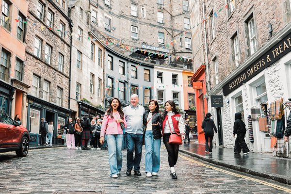 Family of four walking through Edinburgh's wet cobblestone old town street lined with historic stone buildings and shops