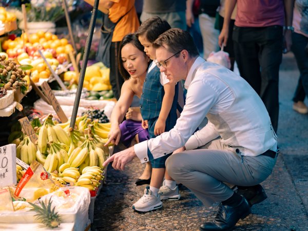 Family exploring colorful tropical fruit market stall in bright daylight, surrounded by bananas and vendors