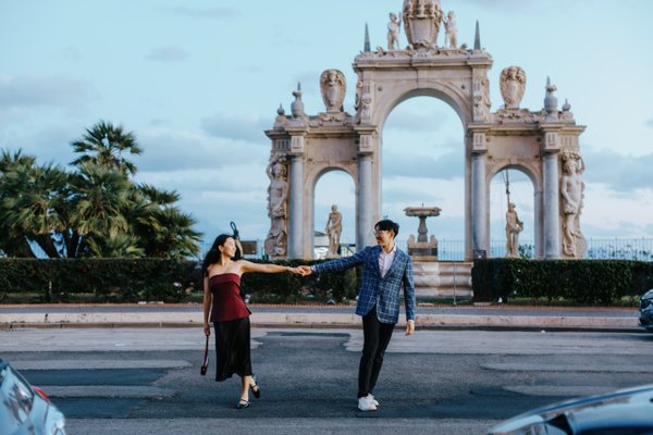 couple holding hands on street with ornate stone archway and palm trees under blue sky