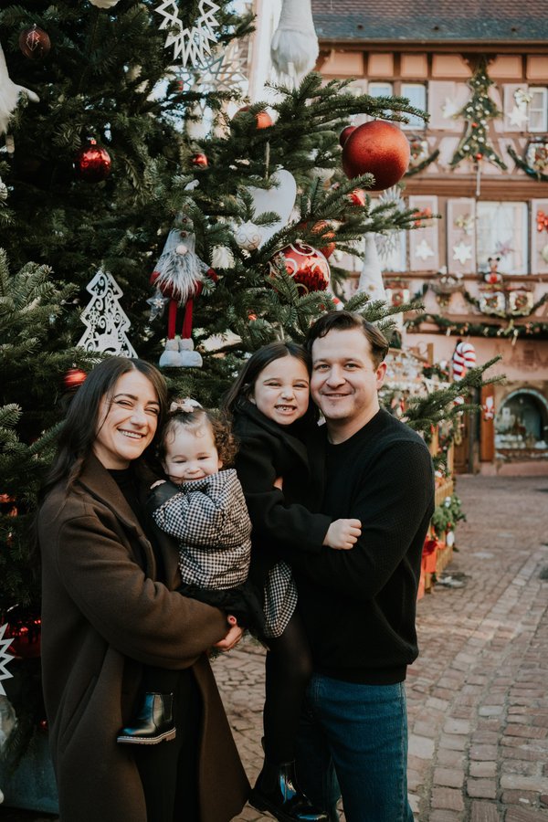 Family of four smiling among decorated Christmas trees in a Colmar street during daytime