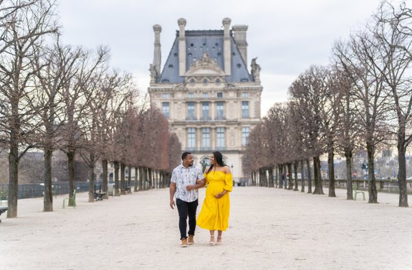 Couple walking arm-in-arm down a tree-lined pathway toward a grand Parisian palace in daylight
