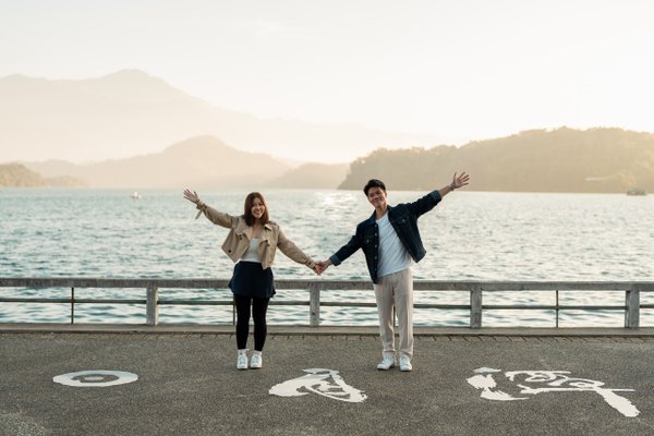 Couple holding hands with arms raised at lakeside promenade, mountains and water in background during overcast day