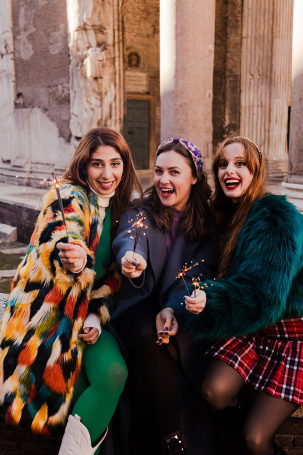 Three women holding sparklers smile at camera in front of weathered Roman columns at golden hour