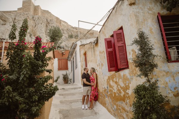 Couple embracing in narrow alley with weathered stone buildings and red shutters, ancient fortress in background