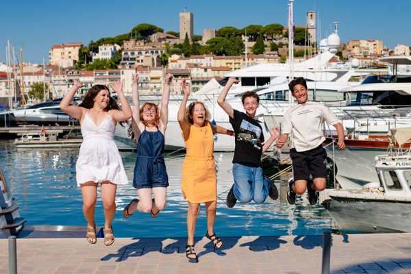 Family of five jumping joyfully on Cannes waterfront with yachts and hilltop castle in bright sunshine
