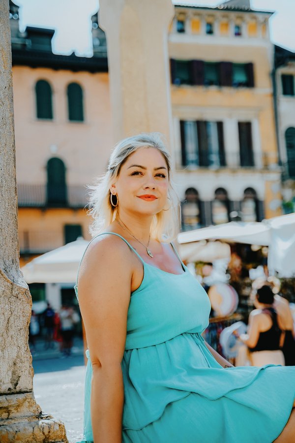 Solo traveler in turquoise dress posing in historic Verona square with golden buildings and market stalls during daytime