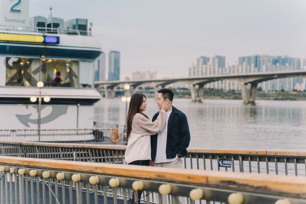 Couple on a waterfront pier with a ferry and Seoul skyline reflected in calm water behind them