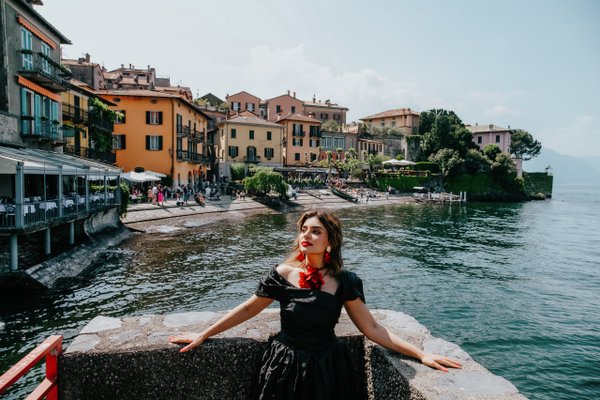 Solo traveler posing at lakeside pier with colorful Italian village and blue water behind, daytime