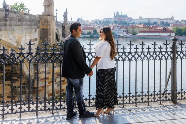 Couple holding hands by ornate railing overlooking Prague's Vltava River and historic cityscape at daytime
