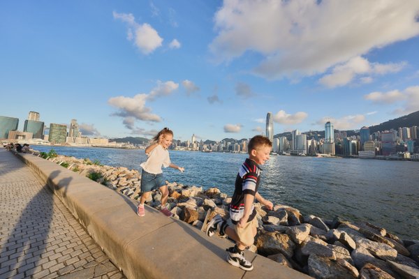 Two children playing on a waterfront promenade with Hong Kong's iconic skyline and Victoria Harbour in bright daylight