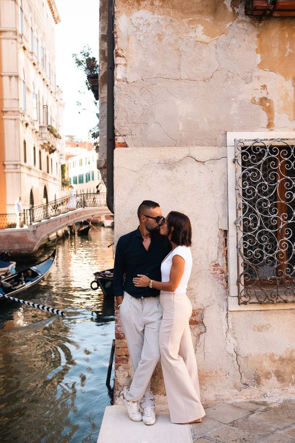 Couple kissing by a weathered canal-side wall in Venice during golden hour, with gondolas and historic buildings nearby