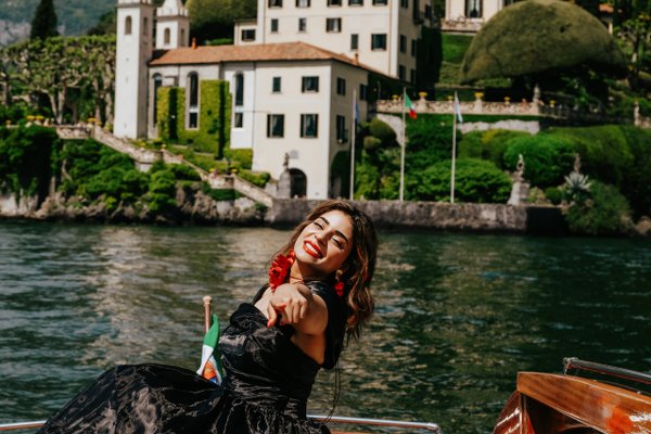 Woman with red earrings on a boat at Lake Como, Italian villa and gardens in bright daylight behind