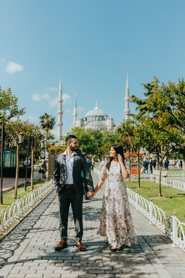 Couple holding hands on a tree-lined promenade with the Blue Mosque and minarets in the background under blue sky