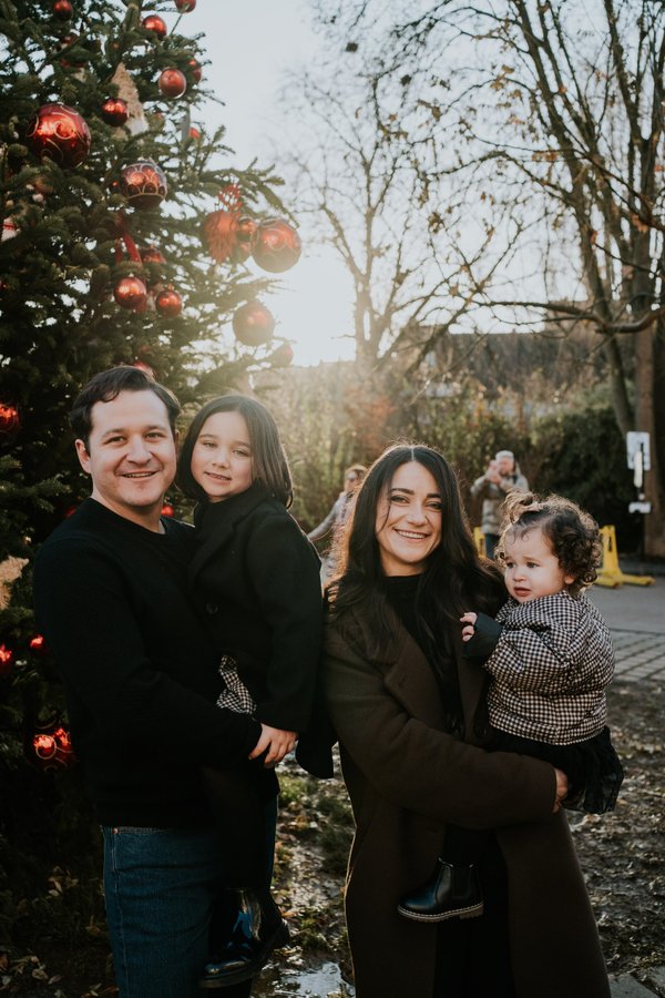 Family of four smiling under red ornaments on decorated evergreen tree at golden hour