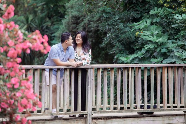 Couple standing on wooden bridge surrounded by lush green foliage and pink flowers in daylight