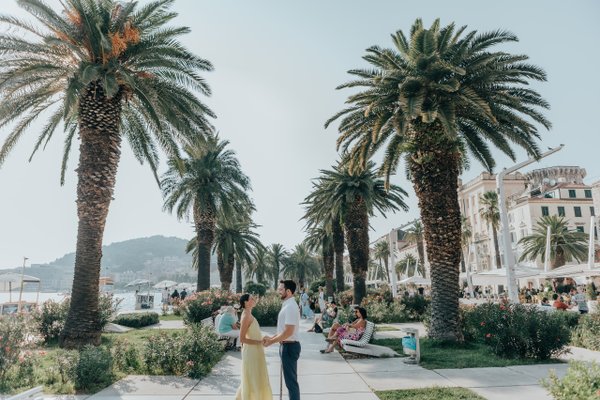 Couple standing among palm trees on a waterfront promenade in Split, with historic buildings and Mediterranean sea beyond