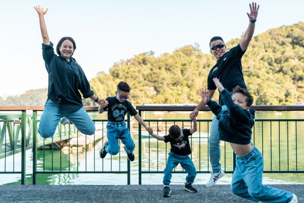 Family of five jumping joyfully on a lakeside deck with forested hills in bright daylight