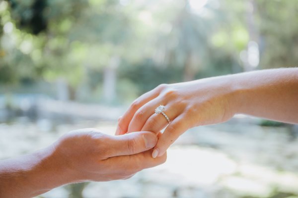 Couple holding hands with diamond engagement ring, lush green garden backdrop in daylight