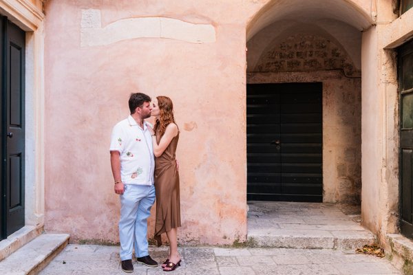 Couple posing against weathered pink stone walls in a historic Mediterranean alleyway during daytime