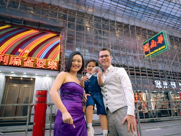 Family posing in Hong Kong's neon-lit street at dusk, with vibrant Chinese signage and urban architecture behind them