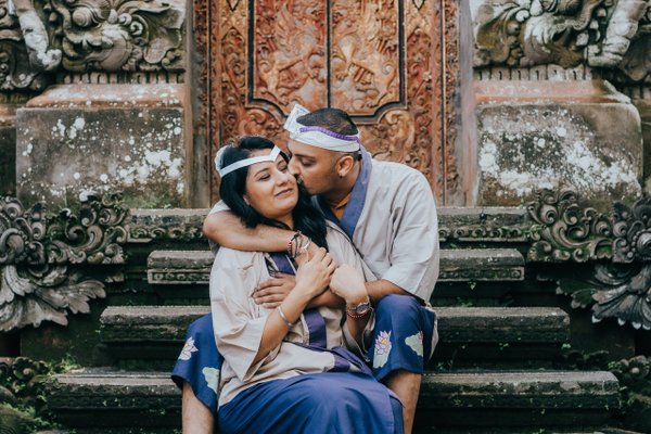 Couple in colorful headbands embrace on weathered stone steps before ornate carved wooden temple doors in Ubud