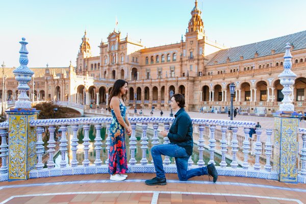 Couple at Plaza de España in Seville during golden hour, man kneeling before woman with ornate tiled bridge and Renaissance palace behind