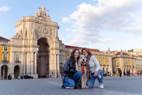 Two women with a terrier dog posing at Praça do Comércio in Lisbon, historic arch and yellow buildings behind them