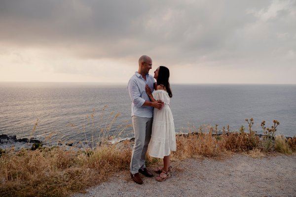 Couple embracing on a clifftop overlooking the sea at golden hour, dry coastal vegetation surrounding them