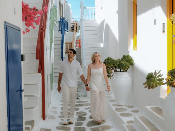Couple holding hands walking through whitewashed Mykonos alleyway with colorful doors and bright sunlight