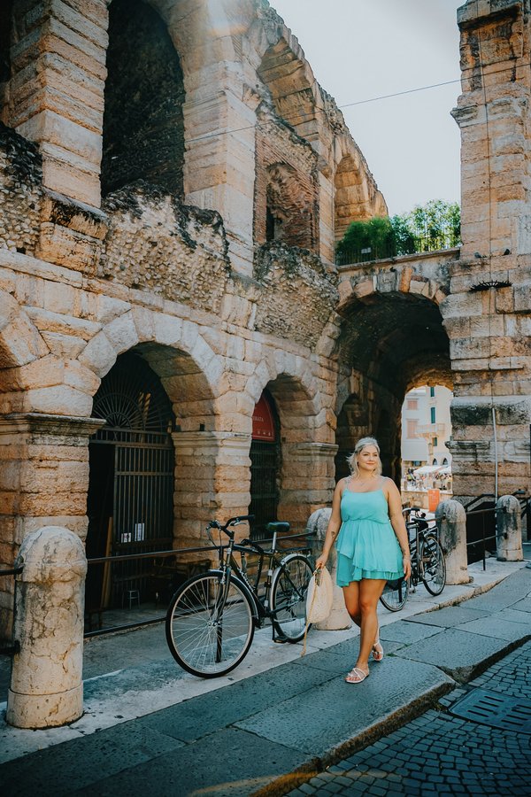 Solo traveler in turquoise dress posing with bicycle near ancient Roman arches in Verona during golden hour