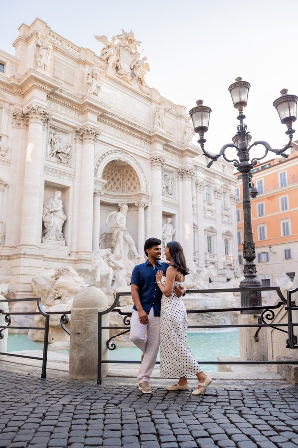 Couple posing at Trevi Fountain in Rome during daytime, with ornate baroque architecture and turquoise water visible behind them