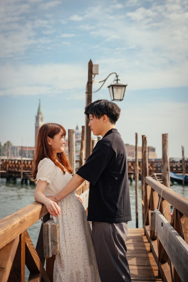 Couple on wooden pier at Punta della Dogana with Venice skyline and moored boats behind in daylight