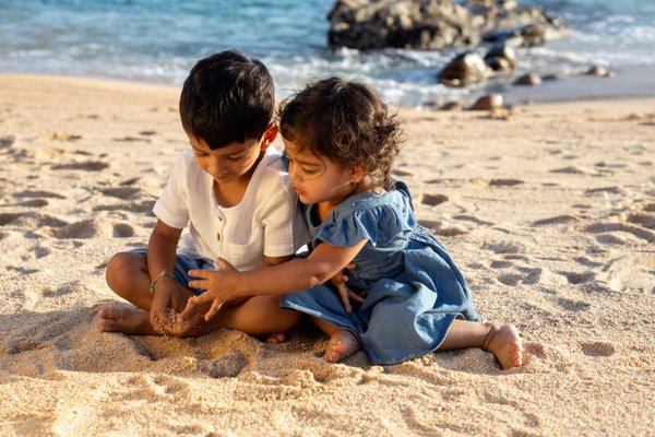 Two young children playing in the sand on a sunny beach with rocky outcrops and turquoise water behind them
